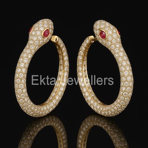 Op de afbeelding: Een paar gouden oorringen in de vorm van slangen. De oorbellen zijn bezet met kleine diamanten en hebben rode edelsteenogen. De tekst "Ekta Jewellers" is op de oorbellen gedrukt.