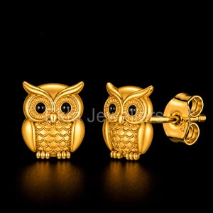 以下が含まれることがあります： 金色のフクロウのスタッドピアス。各イヤリングは、黒い目と質感のある羽を持つ詳細なフクロウのデザインが特徴です。イヤリングは黒い背景に表示され、金属的な光沢と複雑な職人技が強調されています。