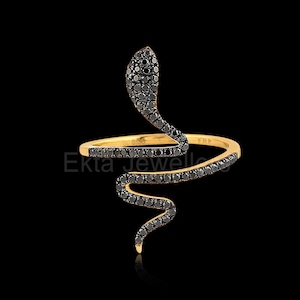 May include: A gold-coloured ring in the shape of a snake, with a head and body that curves around the finger. The snake's body is adorned with black gemstones. The ring is set against a black background.