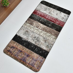 Puede incluir: Una alfombra rectangular tipo pasillero con un diseño de retazos de franjas horizontales en varios colores, incluyendo negro, blanco, gris, rojo, marrón y morado. La alfombra presenta una textura desgastada con patrones desvanecidos.