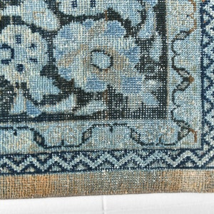 May include: Close-up of the bottom edge of a blue and beige rug with a geometric pattern. The rug has a fringe border.
