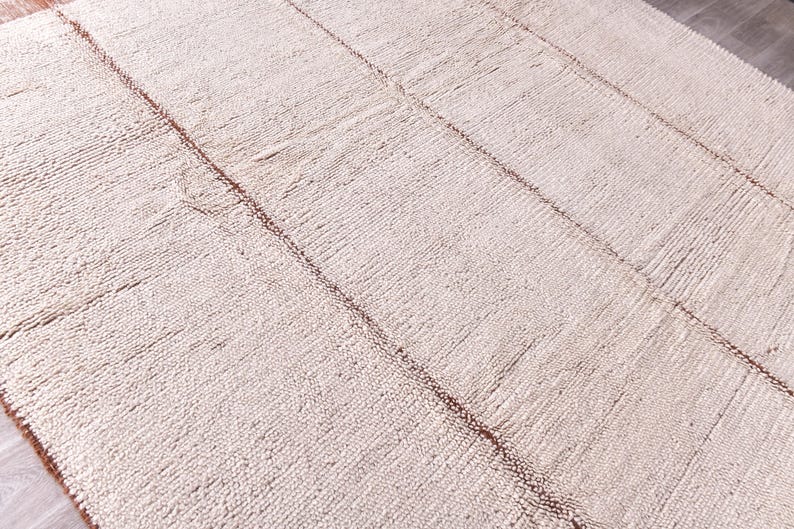 Pu&ograve; includere: Un tappeto rettangolare bianco sporco con una superficie strutturata e linee di accento marroni. Il design del tappeto presenta una serie di linee parallele che corrono lungo la sua lunghezza, creando un motivo sottile. Il tappeto &egrave; realizzato in materiale tessuto.