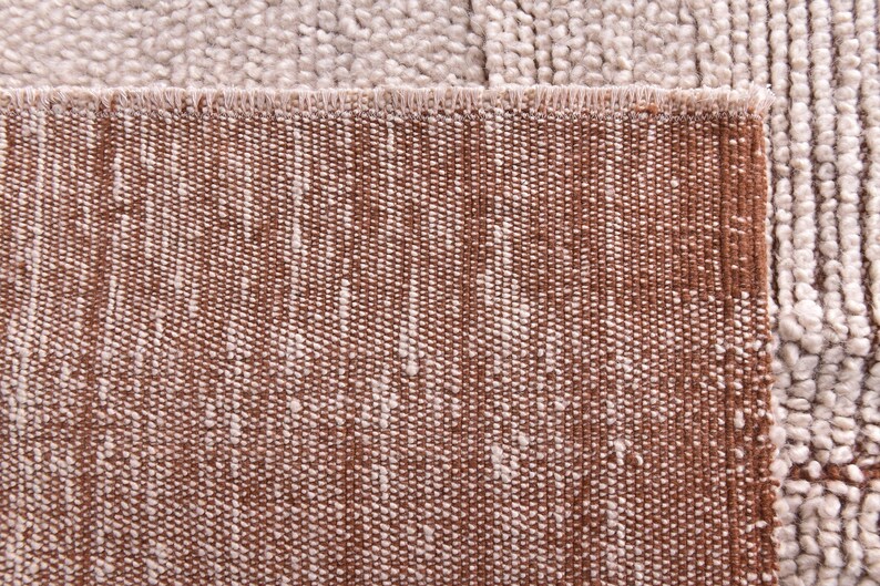 Pu&ograve; includere: Primo piano di un tappeto strutturato con bordo marrone e superficie beige a motivi. Il design del tappeto presenta un mix di trame intrecciate e ad anello, creando una superficie tattile e visivamente interessante. La palette di colori &egrave; neutra, con calde tonalit&agrave; marroni e beige.