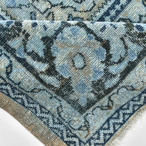 May include: Close-up of a blue and beige patterned rug with a fringed edge. The rug has a floral design with intricate details.