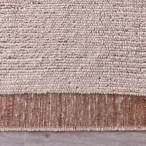 Pu&ograve; includere: Primo piano di un tappeto strutturato con una palette di colori neutri. La parte superiore presenta una trama spessa e nodosa in una tonalit&agrave; beige chiaro. La sezione inferiore ha un bordo tessuto in una tonalit&agrave; marrone pi&ugrave; scura.