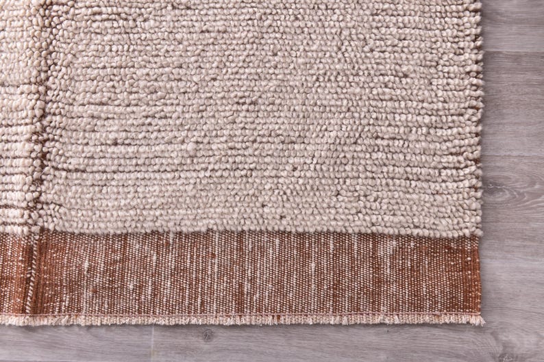 Pu&ograve; includere: Primo piano di un tappeto strutturato con una palette di colori neutri. La parte superiore presenta una trama spessa e nodosa in una tonalit&agrave; beige chiaro. La sezione inferiore ha un bordo tessuto in una tonalit&agrave; marrone pi&ugrave; scura.