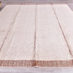 Pu&ograve; includere: Un grande tappeto rettangolare con una superficie strutturata color crema. Il tappeto ha un bordo marrone e strisce verticali marroni. Il tappeto &egrave; realizzato in materiale tessuto ed &egrave; adatto per un soggiorno o una camera da letto.