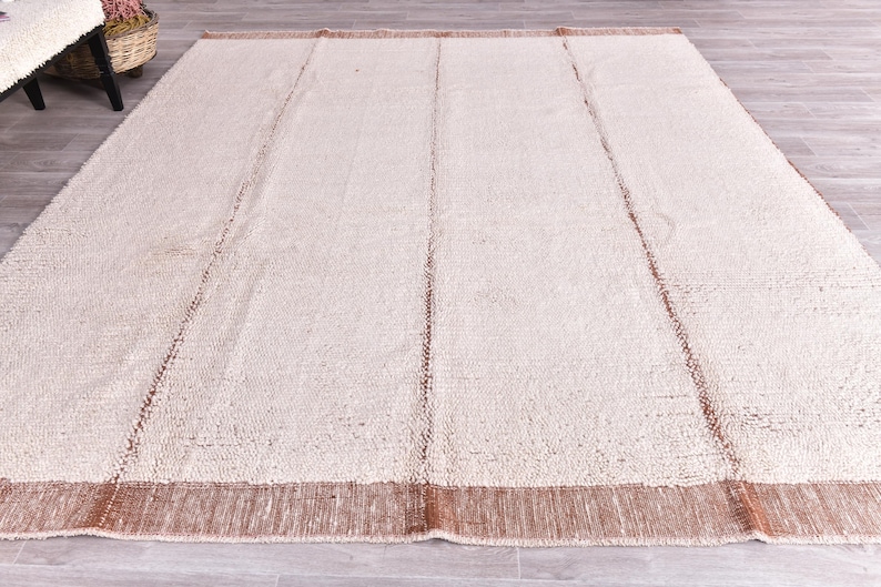 Pu&ograve; includere: Un grande tappeto rettangolare con una superficie strutturata color crema. Il tappeto ha un bordo marrone e strisce verticali marroni. Il tappeto &egrave; realizzato in materiale tessuto ed &egrave; adatto per un soggiorno o una camera da letto.