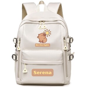 May include: A light beige backpack with multiple compartments and adjustable straps. It features a cartoon illustration of a bear and sun, with the text "Be the light" and "Serena". The backpack has a zippered front pocket and side pockets.