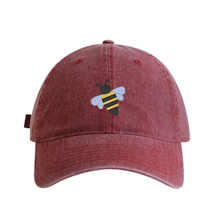 May include: A maroon baseball cap with a small embroidered bee design on the front. The bee is black, yellow, and light blue. The cap has a curved brim and a fabric strap closure.