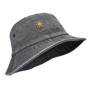 Op de afbeelding: Een antracietgrijze bucket hat met een verwassen, verweerde textuur. De hoed is voorzien van een klein, geel geborduurd zonnepictogram in het midden van het voorpaneel. Het heeft een neerwaarts hellende rand en een gestikte band rond de kroon.