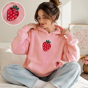 May include: A light pink hooded sweatshirt featuring a centered embroidered strawberry design with a red body, white seeds, and green leaves. The hoodie includes a front kangaroo pocket and long sleeves, with a circular inset showing the detail.