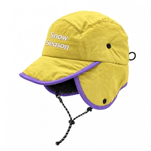 May include: A mustard yellow winter hat with ear flaps, featuring the white embroidered text Snow Season on the front. The hat has a purple trim, a black adjustable chin strap with a toggle, and a dark gray textured lining inside.