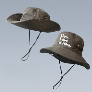 Puede incluir: Dos sombreros de cubo verdes con correas de barbilla negras. Un sombrero tiene el texto "Live, Love & Hike." bordado en la parte delantera.