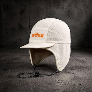 May include: An off-white cap with ear flaps and a short brim. The word arthur is embroidered in orange on the front panel. A black cord with a plastic toggle is attached to the bottom of the ear flaps, allowing for adjustment under the chin.