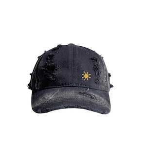 May include: A distressed black baseball cap with a yellow sun embroidered on the front. The cap has a worn, faded appearance with intentional rips and tears for a vintage look. The brim is curved.