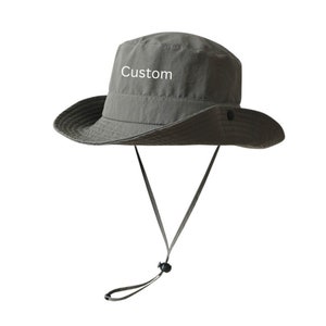 以下が含まれることがあります： 黒い紐が付いた、灰色のつば広の帽子。帽子には「Custom」という文字が刺繍されています。