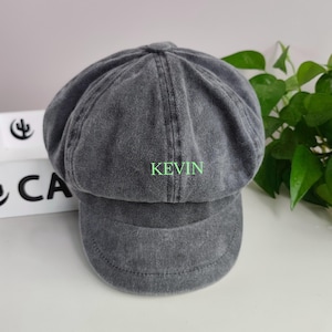 Może przedstawiać: Ciemnoszara czapka typu gazeciarz o spranym, postarzanym wyglądzie. Z przodu czapki wyhaftowano imię KEVIN jaskrawozielonymi, wielkimi literami. Nakrycie głowy posiada usztywnianą górę z wieloma panelami oraz krótki, sztywny daszek z przodu.