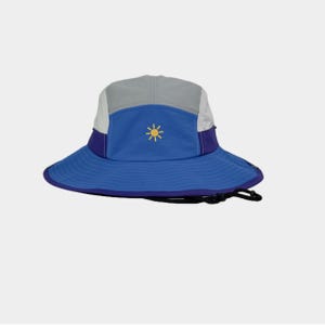 Op de afbeelding: Een bucket hat met meerdere panelen, voorzien van een blauwe kroon en rand, lichtgrijze zijpanelen en een donkerpaarse band. Een klein geel zonnepictogram is in het midden aan de voorzijde geborduurd. De hoed heeft een zwarte kinband.