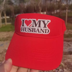 May include: Red visor with a white sticker that says "I love my husband" with a red heart.