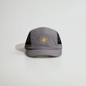 May include: A grey five-panel cap featuring a yellow sun icon embroidered on the front center. The hat is constructed with a smooth, crinkled fabric crown and black mesh side panels, finished with a solid grey brim and a structured front panel.