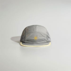 Peut inclure: Une casquette à cinq panneaux gris clair avec une icône de soleil brodée en jaune sur le devant. Les panneaux latéraux sont en maille grise et la visière plate est bordée d'une finition contrastante de couleur crème clair.