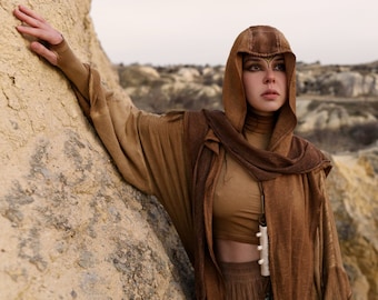 May include: A person wearing a hooded brown robe with long sleeves, a matching scarf, and a crop top. The outfit includes a decorative pendant. The person is leaning against a rock formation, with a desert landscape in the background.