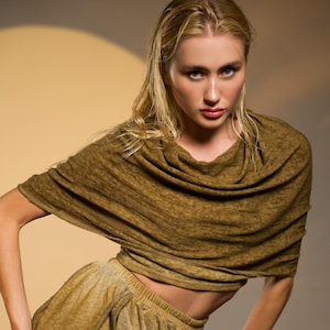 May include: A woman wearing a two-piece olive green outfit. The top is a draped, cowl-neck style, and the pants are loose-fitting with a drawstring. The fabric appears soft and textured, with a slight sheen. The background is a neutral beige.