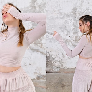May include: A woman is wearing a light pink, long-sleeved, cropped top and matching pants. The top has a high neck and the pants have a wide leg.