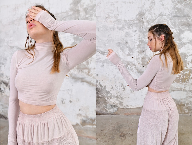 May include: A woman is wearing a light pink, long-sleeved, cropped top and matching pants. The top has a high neck and the pants have a wide leg.