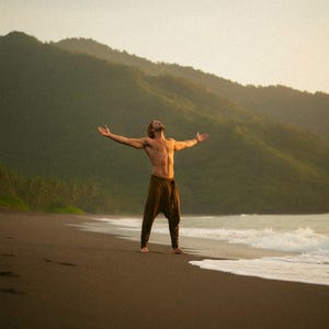 May include: A person stands on a dark beach with arms outstretched, facing the ocean. They wear brown pants. The background features green mountains and a light sky, creating a serene outdoor scene.