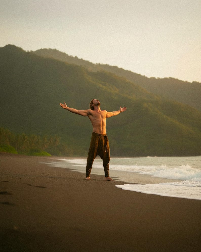 May include: A person stands on a dark beach with arms outstretched, facing the ocean. They wear brown pants. The background features green mountains and a light sky, creating a serene outdoor scene.