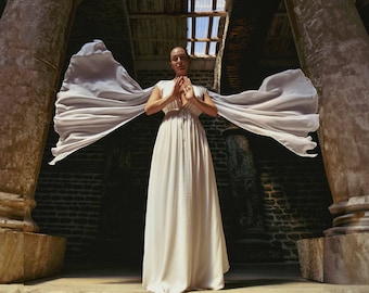 May include: A woman in a white dress stands with hands clasped in front of her chest. A long, flowing white fabric extends from her shoulders, creating a dramatic effect. The setting is an ancient stone structure with large columns and a brick wall.