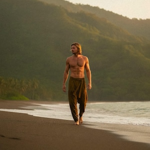 May include: A person wearing brown harem pants walks barefoot on a beach. The person is shirtless, with long blond hair. The background features a mountain range and the ocean. The pants have a wrap-around design.