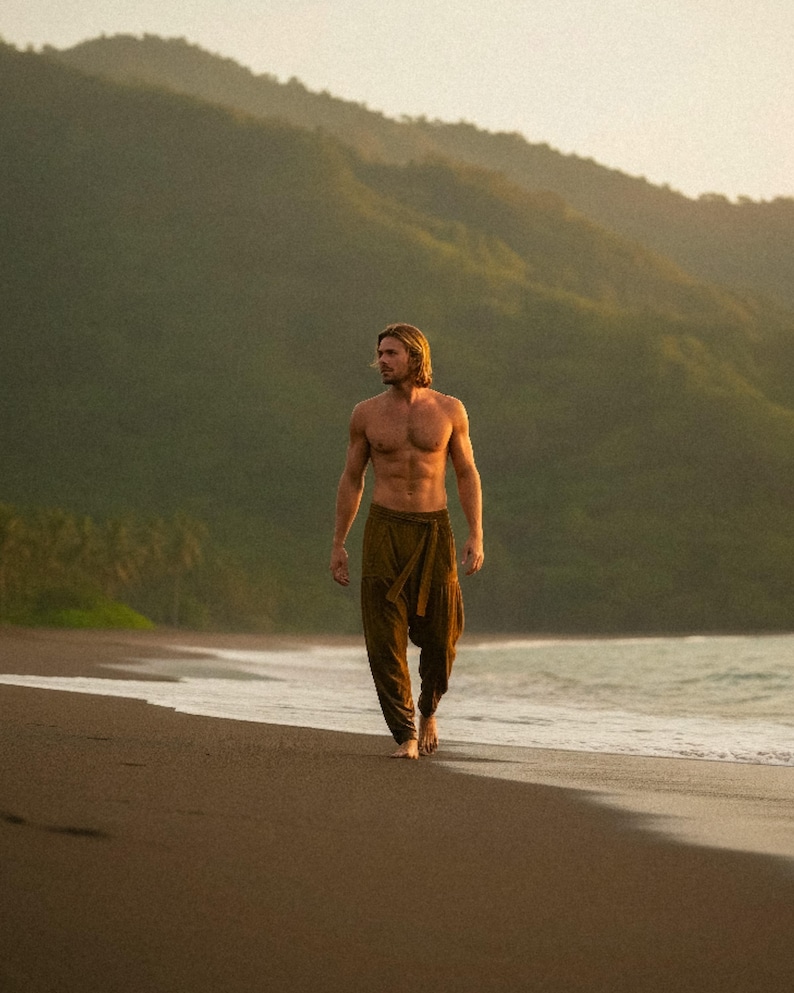May include: A person wearing brown harem pants walks barefoot on a beach. The person is shirtless, with long blond hair. The background features a mountain range and the ocean. The pants have a wrap-around design.