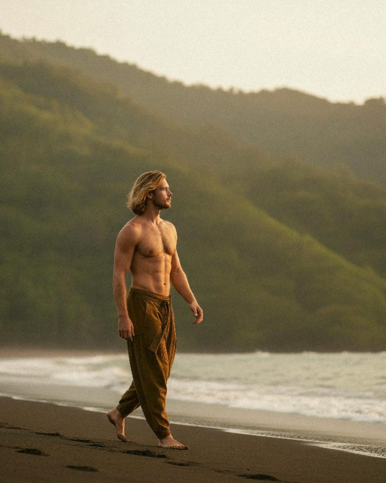 May include: A person with long blond hair and brown pants walks barefoot on a beach. The person is shirtless, with a mountain range in the background. The sky is overcast.