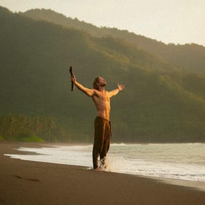 May include: A person stands on a black sand beach with arms outstretched, holding a wooden stick. The person is wearing brown pants. The background features a mountain range and the ocean under a hazy sky.