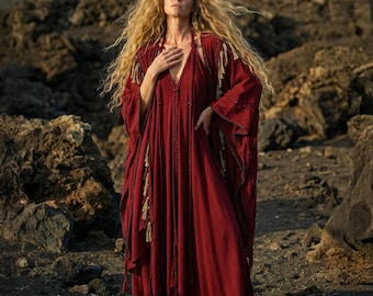May include: A flowing, deep red dress with long sleeves and a plunging neckline. The dress features decorative tassels and is worn outdoors against a backdrop of rocky terrain. The model has long, wavy blonde hair.