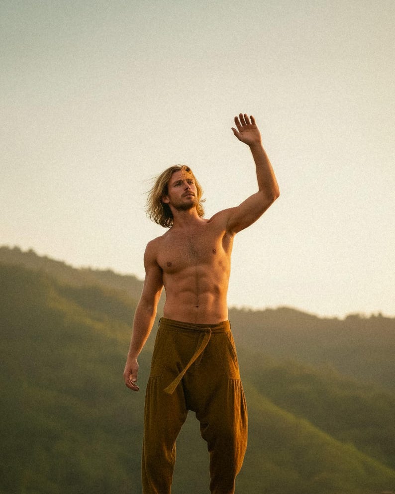 May include: A person with long blonde hair wearing olive green harem pants stands outdoors with their arm raised. The person is shirtless, with a mountainous landscape in the background. The lighting suggests a sunset or sunrise.