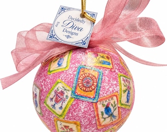 May include: A spherical pink ornament with a textured surface featuring various playing card illustrations, including a Joker and floral designs. A sheer pink ribbon is tied at the top, and a square tag reads Decidedly Diva Designs.