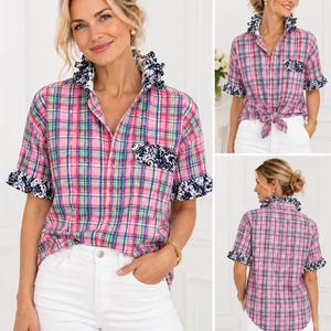 May include: A short-sleeved button-down shirt featuring a pink, green, and white plaid pattern. The shirt includes a dark blue and white floral print on the ruffled collar, sleeve cuffs, and chest pocket. The hem is tied at the waist in the front.