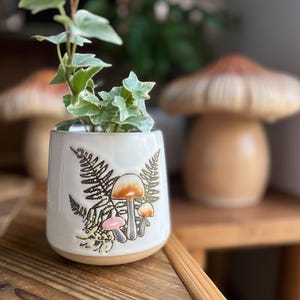 May include: A small, off-white ceramic planter with a botanical design featuring ferns and mushrooms. The planter holds a small plant with green and white leaves. The background includes a wooden table and a mushroom-shaped decorative item.