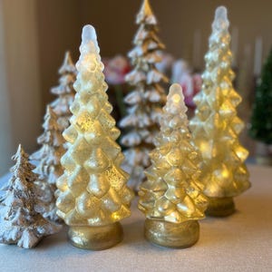 May include: A collection of decorative Christmas tree figurines in various sizes. The trees are primarily gold and silver, with textured surfaces and a frosted appearance. Some trees have a clear, pointed top, and all are set on a neutral surface.