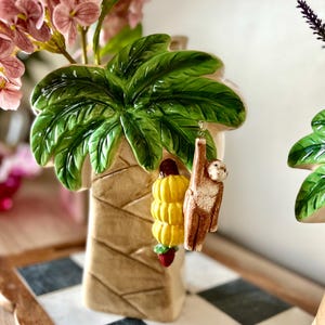 May include: A ceramic palm tree vase with a green palm leaf top and a brown trunk. A small monkey hangs from a bunch of yellow bananas. Pink flowers are in the vase. The vase sits on a black and white checkered surface.
