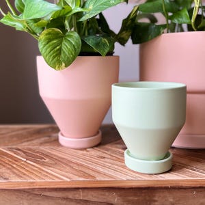 May include: Three ceramic plant pots in shades of pink and green. The largest pot is a light pink, the medium pot is a two-tone pink, and the smallest pot is a pale green. All pots have matching saucers.