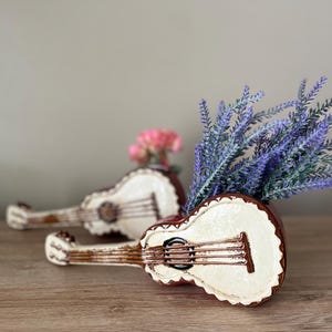 May include: Two ceramic guitar-shaped planters, one in focus, filled with lavender and pink flowers. The guitars are brown and cream-colored with decorative details. The background is a neutral color, and the planters sit on a wooden surface.