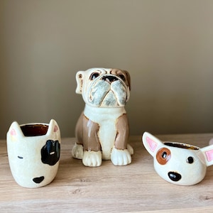 May include: Three ceramic dog-shaped planters. The central planter is a bulldog in brown and white. Two smaller planters are shaped like dog faces, one with a black patch and the other with a brown ear spot. The planters are on a wooden surface.