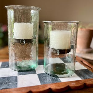May include: Two clear glass candle holders with textured surfaces. Each holder contains a white pillar candle held in place by a dark metal band. The holders are different sizes and sit on a black and white checkered tray.