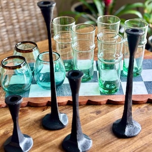 May include: A collection of three black metal candle holders with tapered stems and circular bases, arranged alongside several clear green glass tumblers and rounded glasses, all positioned on a checkered board surface.
