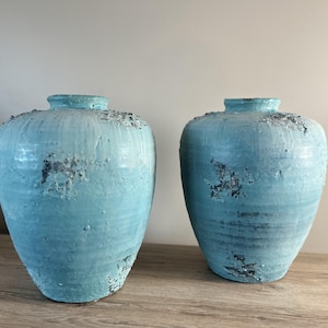 May include: Two turquoise ceramic vases with a distressed finish. The vases have a rounded shape with a narrow neck and a textured surface. The vases are displayed on a wooden surface.